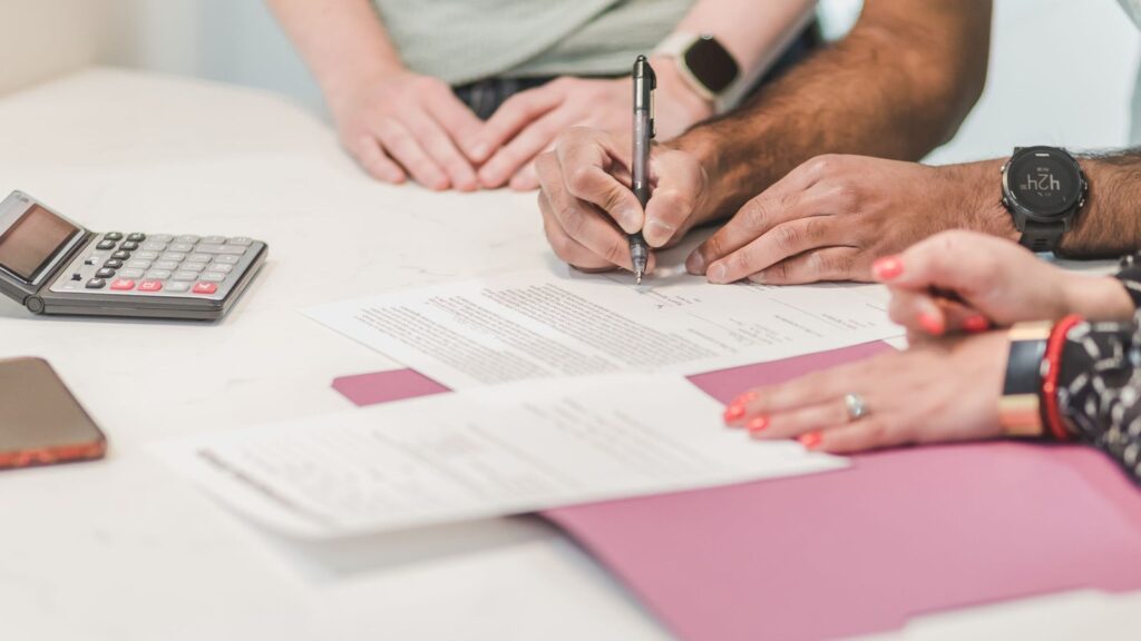 Business buyer signing loan documents at a table with a lender, calculator and paperwork nearby