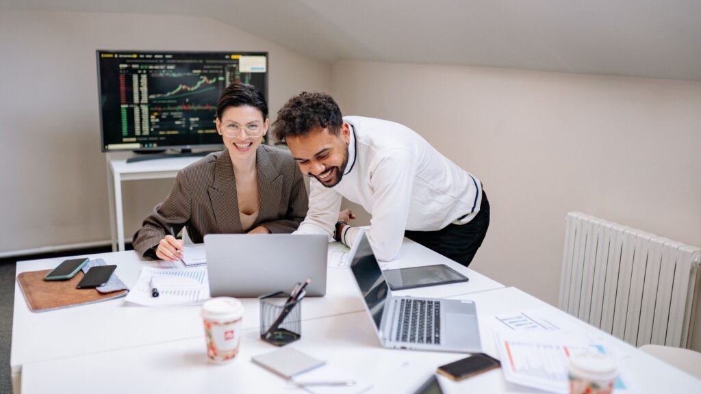 Two colleagues smiling and reviewing files together on laptops in a bright office