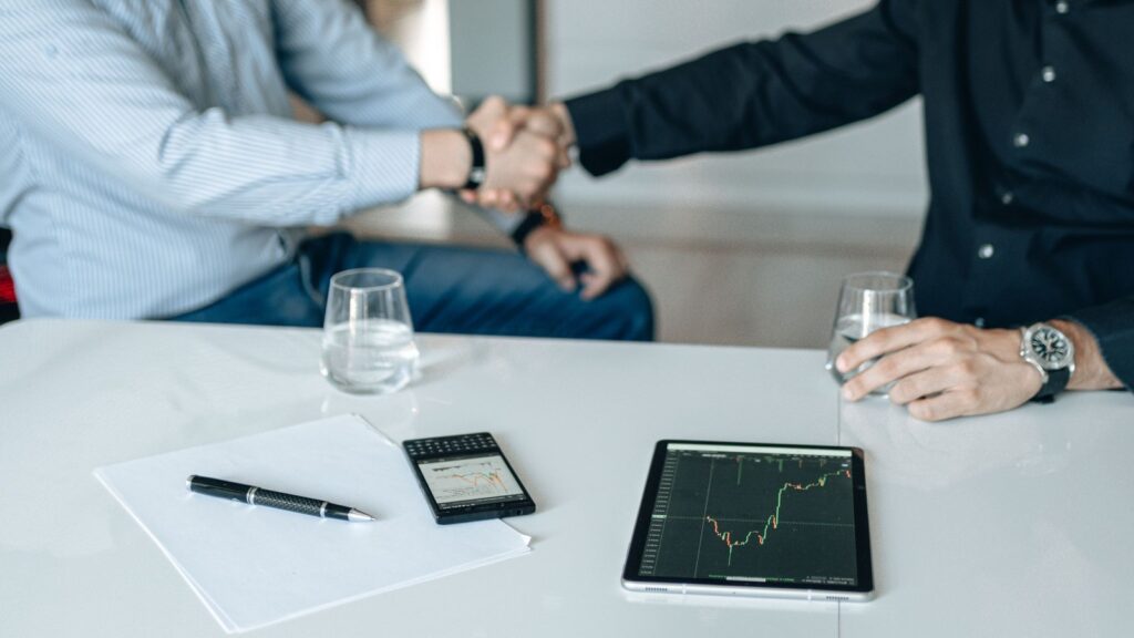 Investors shaking hands across a meeting table with a tablet showing a financial chart
