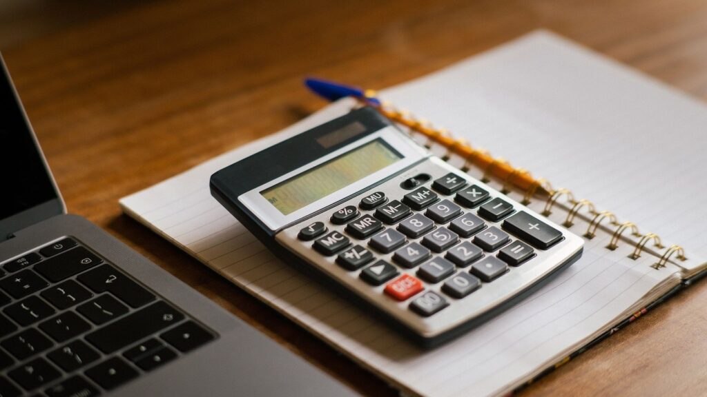 Calculator resting on a notebook next to an open laptop on a wooden desk