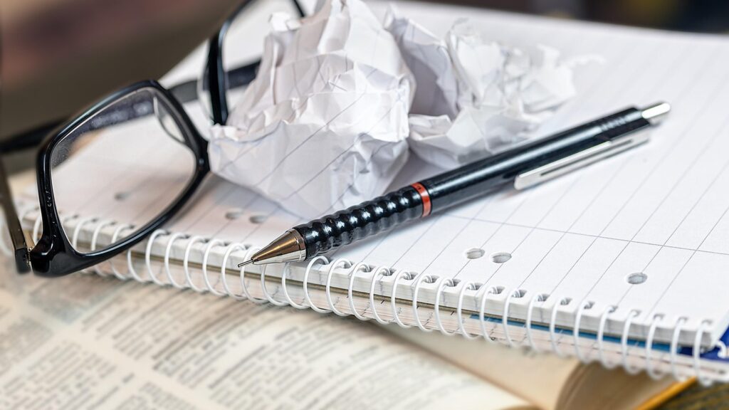 Glasses, a pen and a spiral notebook with crumpled paper on a desk