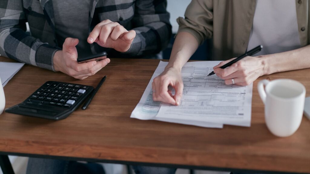 Two people completing tax forms at a wooden desk with a calculator and smartphone