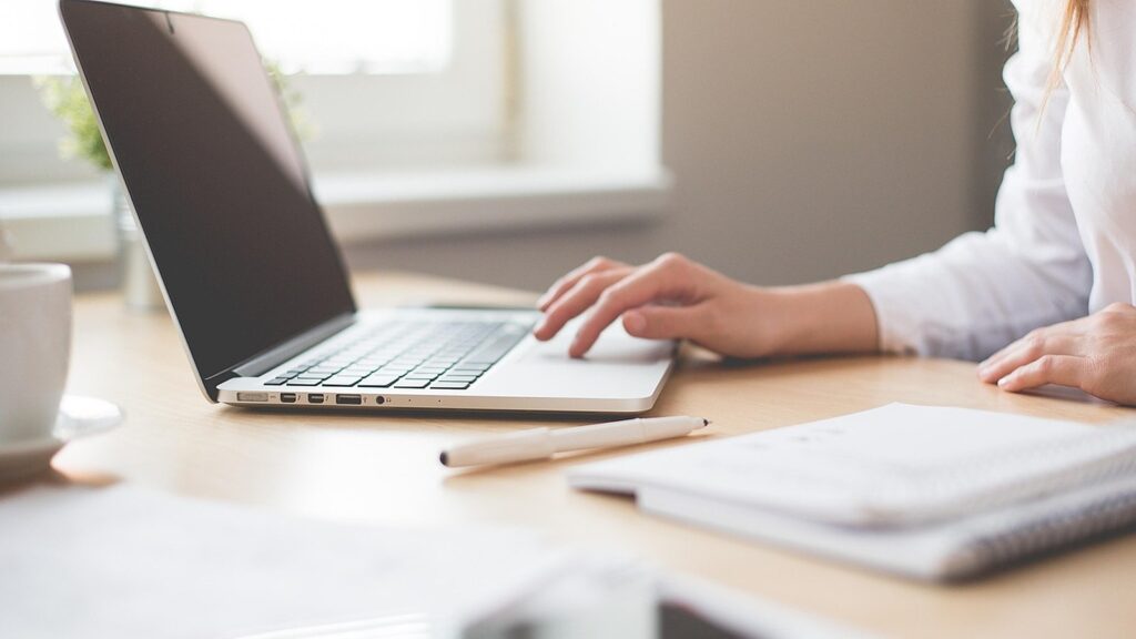 Person typing on a laptop at a bright desk with a notebook beside it