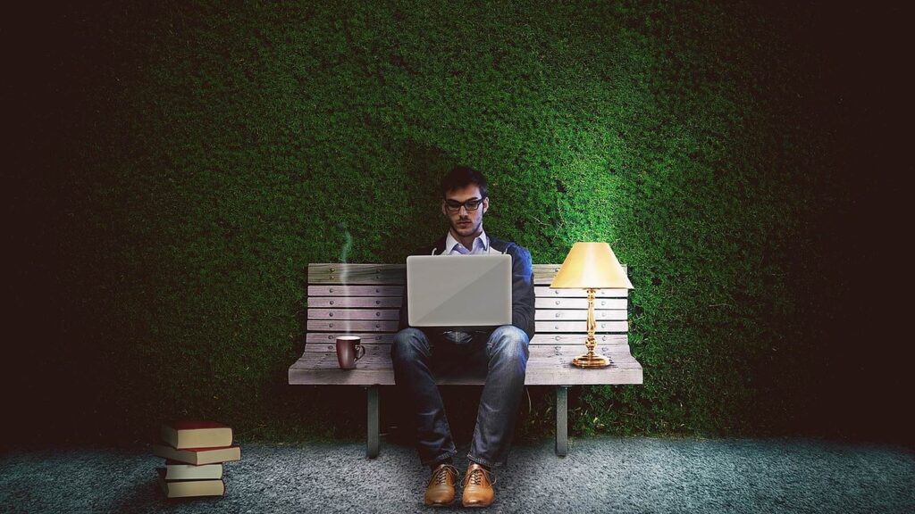 Man working on a laptop on a bench at night beside a lamp and a stack of books
