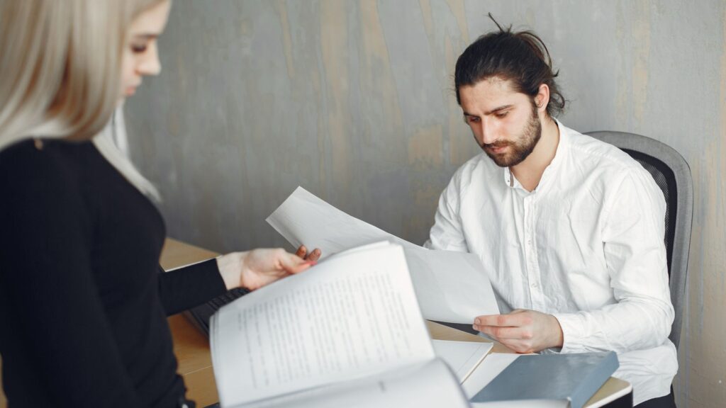 Two professionals in a modern office reviewing printed documents together, one seated at a desk and one standing