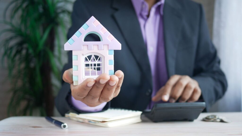 Real estate professional in a suit holding a model house while using a calculator at a desk