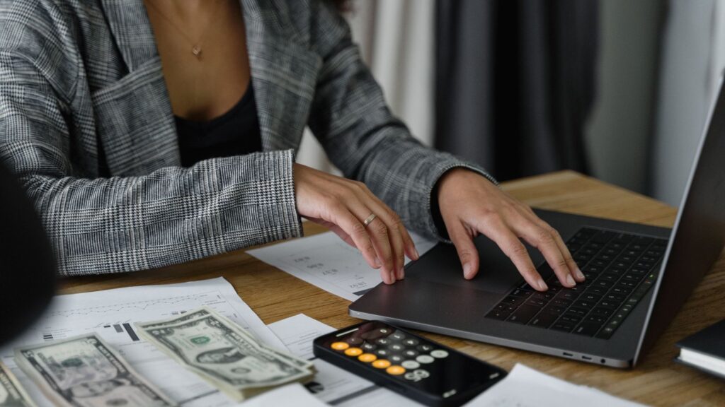 Business owner working on a laptop beside cash, a calculator and payroll paperwork on a desk