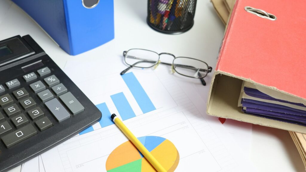 Desk with a calculator, pencil, eyeglasses, printed bar and pie charts, and colored binders of financial paperwork